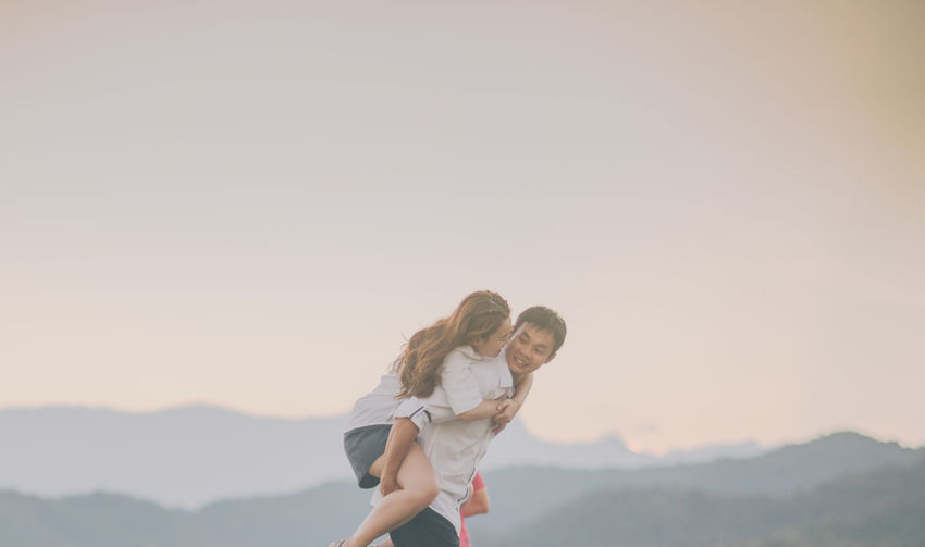 Man carrying woman while standing against sky during sunset