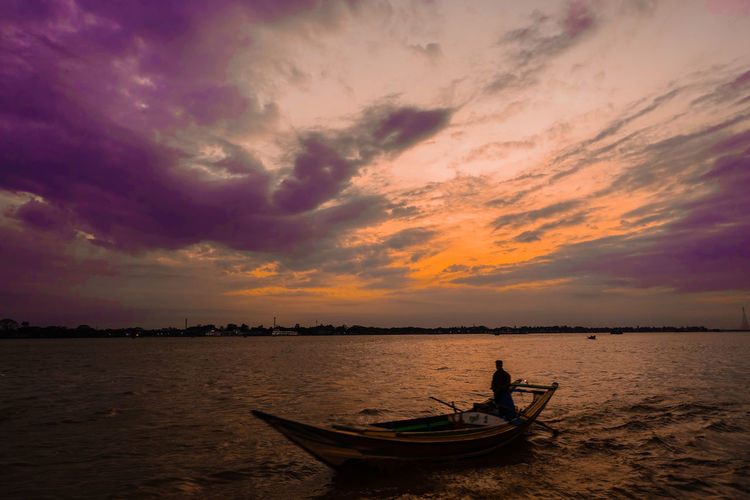 Silhouette man on boat in river against cloudy sky during sunset