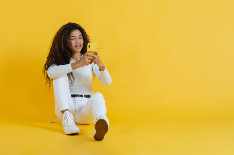Young woman using mobile phone while standing against yellow background