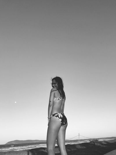 Low angle view of smiling woman in bikini standing at beach against clear sky