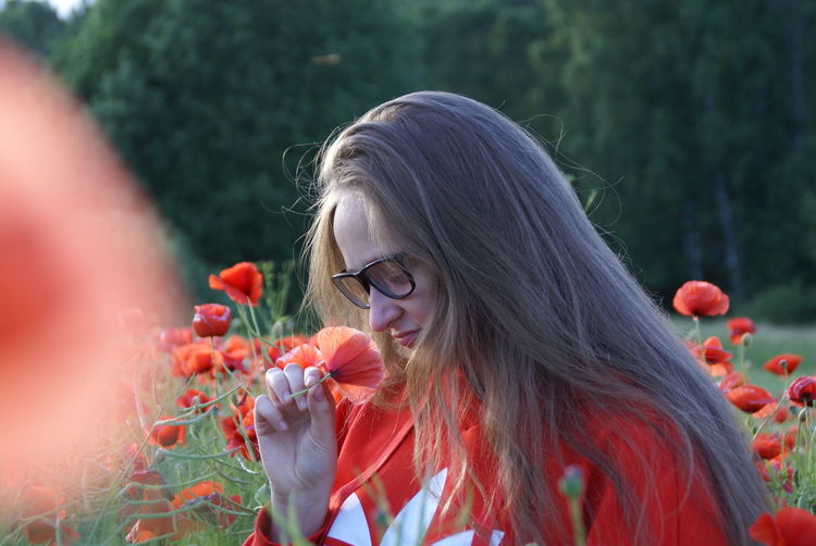 Young woman in eyeglasses smelling poppy while sitting on field
