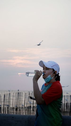 Side view of woman standing drinking water against sky