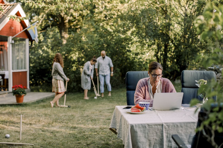Woman using laptop while family playing in background at yard