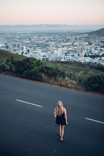 Rear view of woman standing on road by cityscape against clear sky during sunset