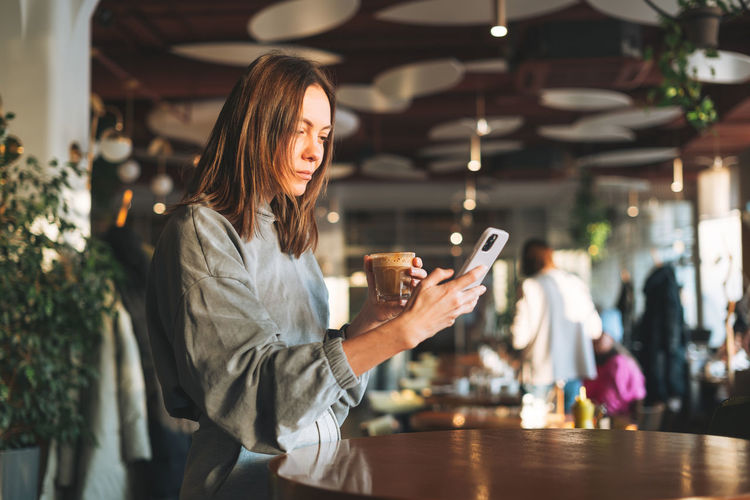 Young woman wearing casual longsleeve with mobile phone in hands with cup of coffee sitting in cafe