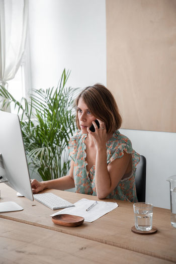 Concentrated female freelancer having phone conversation while surfing computer during online work at table in light home office