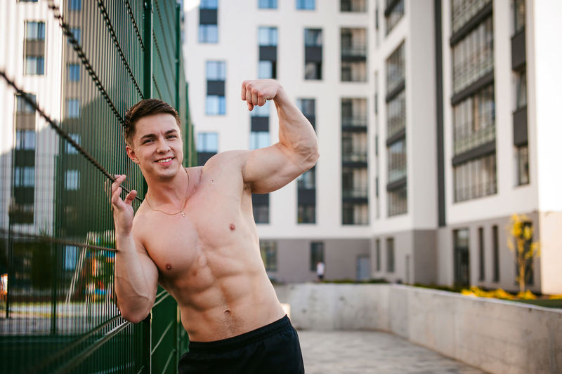 Portrait of muscular man standing by fence at park
