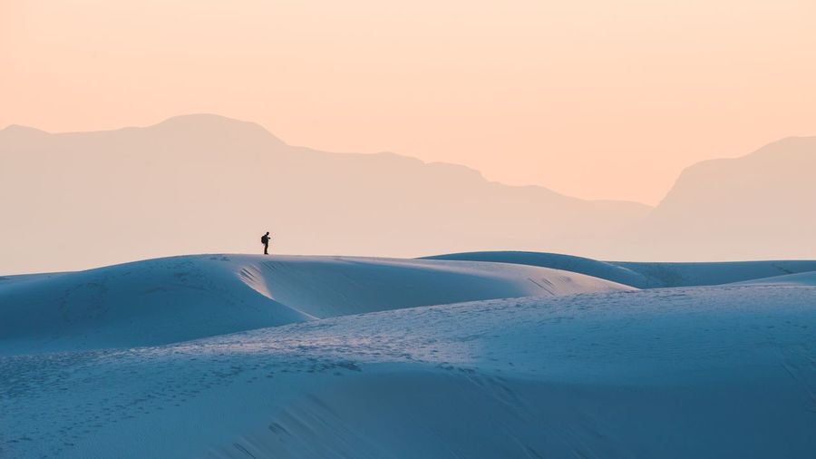 Distant view of man standing on sand dunes against sky during sunset