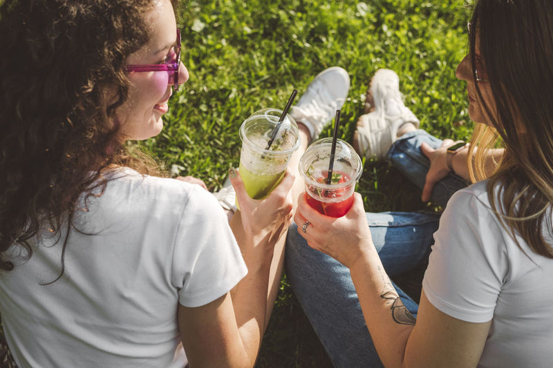 Women toasting fresh lemonade in disposable cups at park on sunny day