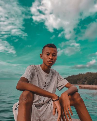 Young man sitting by sea against sky