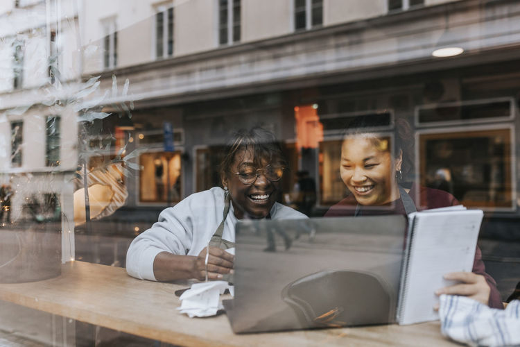 Happy female coffee shop owners discussing over laptop seen through window