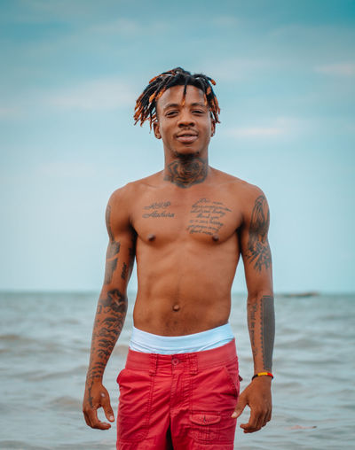 Portrait of young man with tattoos standing at beach against sky