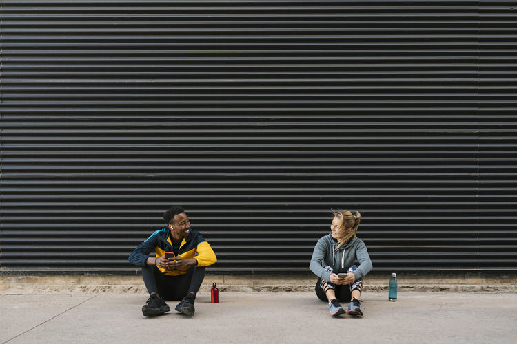 Smiling male and female athlete with mobile phone looking at each other while sitting against wall