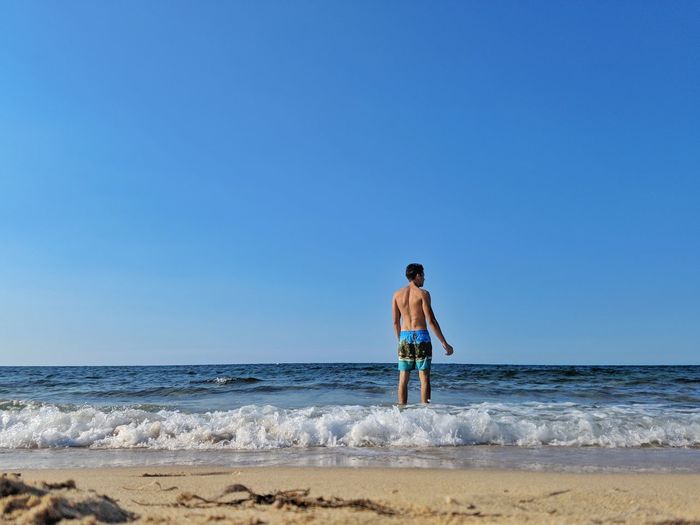 Rear view of shirtless man standing on shore at beach against clear sky