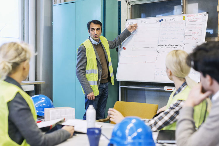 Mature male worker explaining plan on whiteboard to colleagues in factory