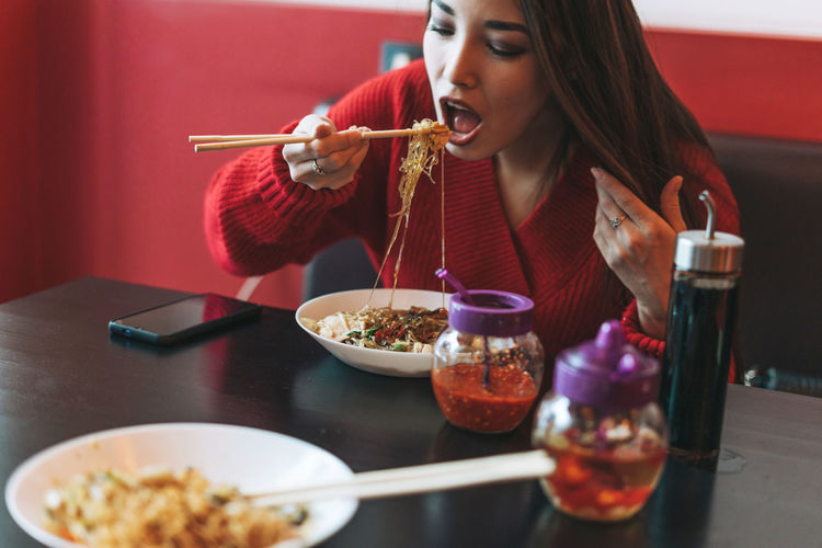 Beautiful young asian woman in red clothes eating noodles with chopsticks in chinese restaurant