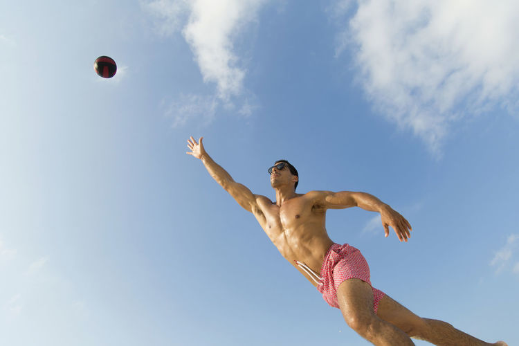 Shirtless man playing volleyball against sky