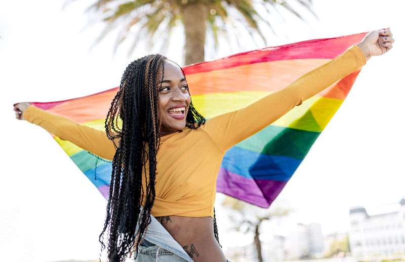 Low angle of proud african american female standing with fluttering lgbt rainbow flag on street and cheerfully looking away