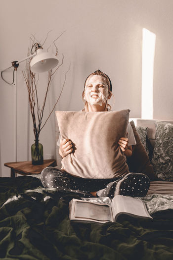 Woman with face mask holding cushion while sitting on bed at home