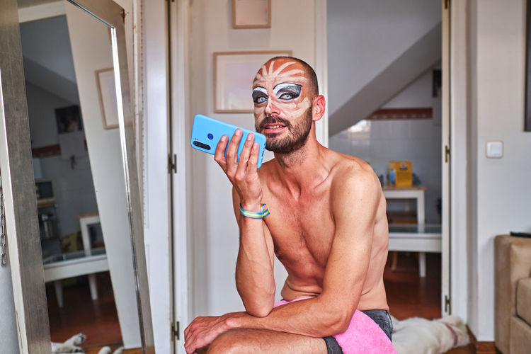 Eccentric shirtless bearded male with drag queen makeup and pride bracelet recording voice message on blue cellphone while sitting near mirror at home