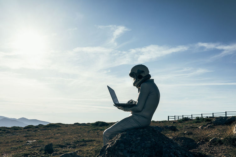 Full body male astronaut in spacesuit browsing data on netbook while sitting on rock outside station with rocket shaped antennas