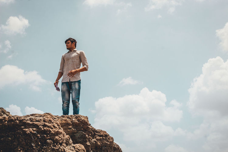 Low angle view of young man standing on rock against sky