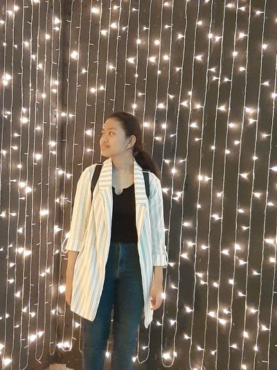 Young woman looking away while standing against illuminated wall