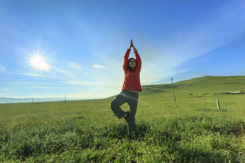 Young woman doing yoga on grassy landscape against sky during sunny day