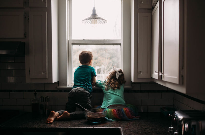 Young boy and girl staring out kitchen window on rainy day