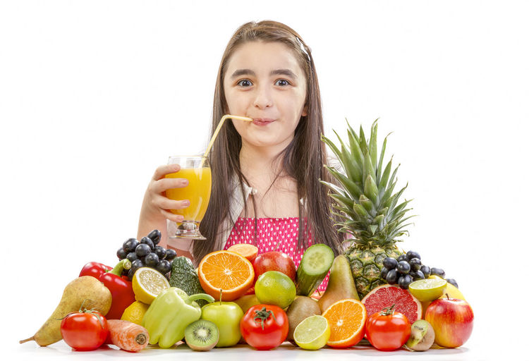 Portrait of teenage girl drinking juice while sitting with fruits on table against white background