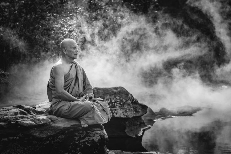 Full length of monk meditating while sitting on rock by river