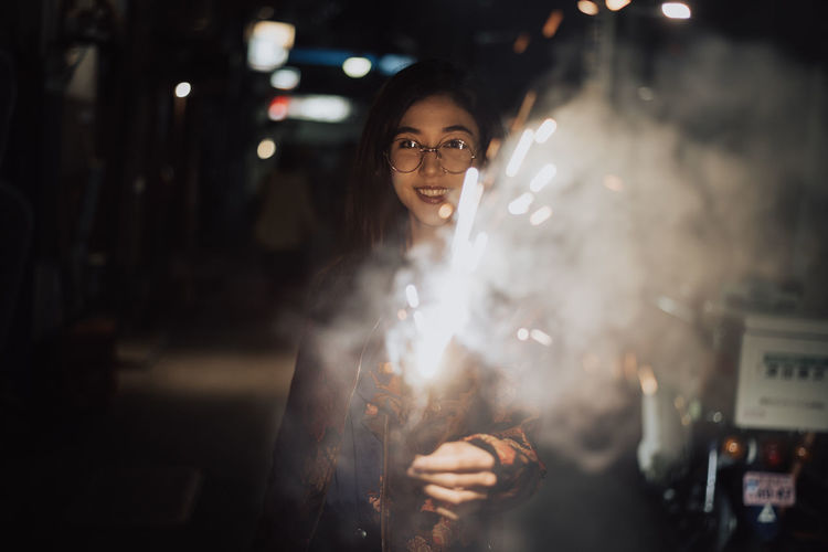 Portrait of smiling woman holding sparkler standing at night outdoors