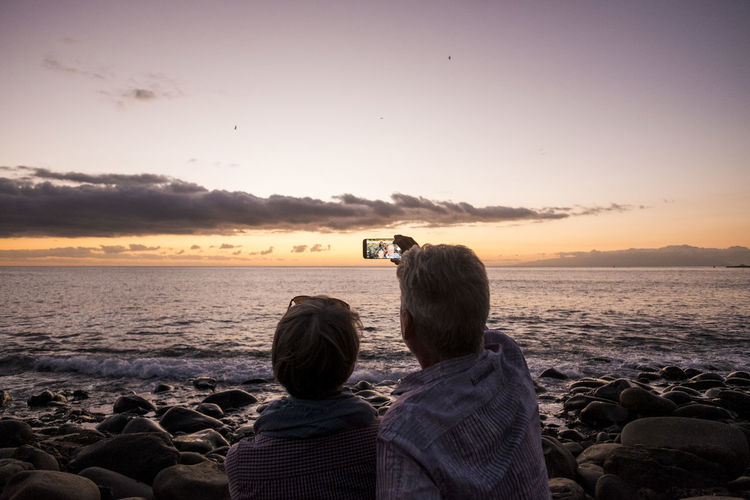 Rear view of man with woman taking selfie from mobile phone at beach