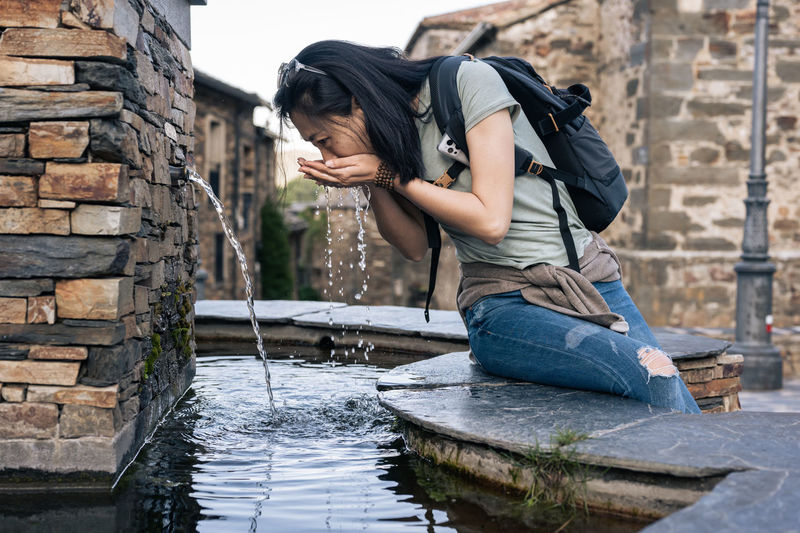 Asian female tourist with backpack drinking water from fountain on street with aged stone buildings in old town during trip in valverde de los arroyos