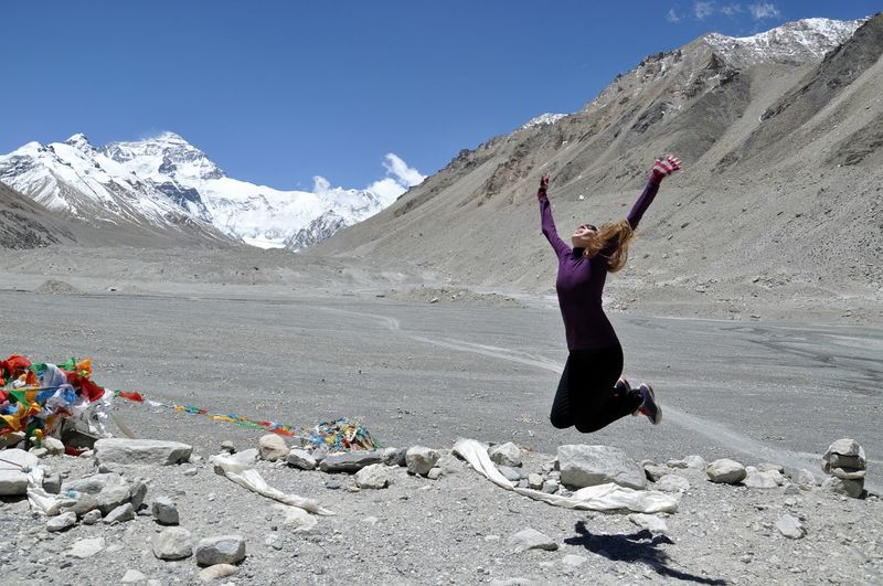 Full length of woman jumping in mid-air over rocks against snowcapped mountains
