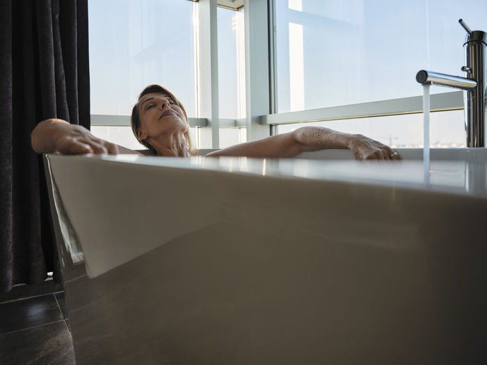 Relaxed senior woman taking bath in bathtub against window at luxury hotel room