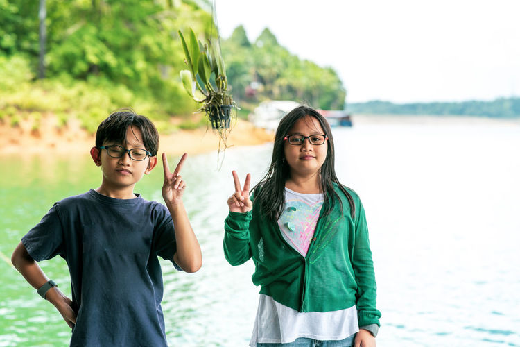 Siblings, boy and girl wearing glasses showing peace sign while travelling to the lake.