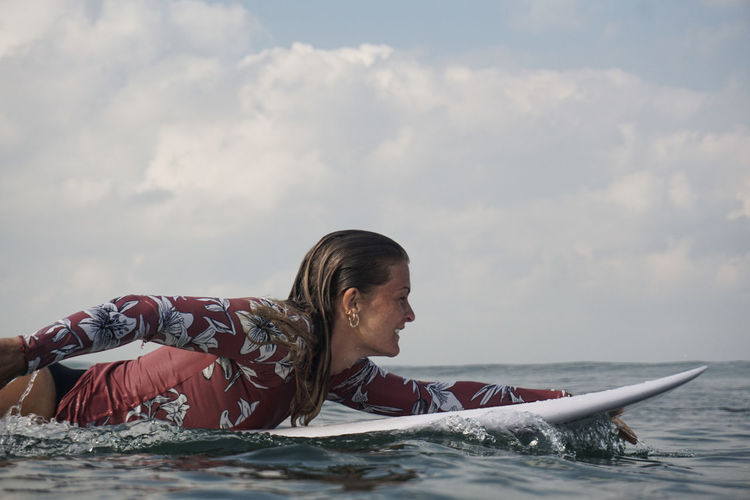Side view of woman lying on surfboard in sea against sky