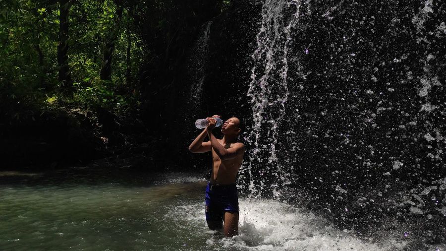 Shirtless young man drinking water from bottle while standing by waterfall