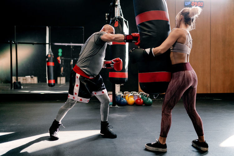 Side view of strong female athlete in boxing gloves holding punching bag while man practicing punches during workout together in modern boxing gym