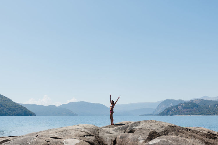 Woman with arms raised in bikini standing at lakeshore against clear sky during sunny day