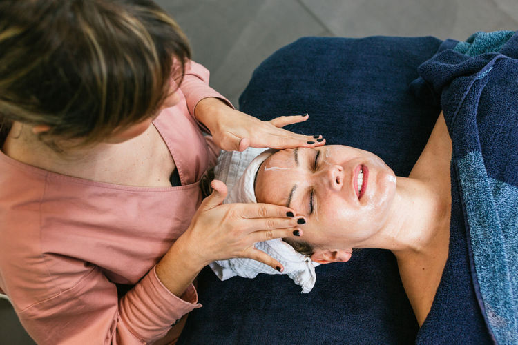 From above crop unrecognizable cosmetician applying facial cleanser on face of female client during skin care treatment in beauty salon