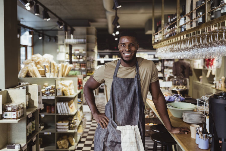 Portrait of smiling male owner with hand on hip standing in delicatessen shop
