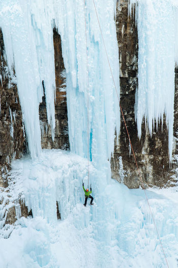 An ice climber dangles from a frozen waterfall scaling challenging snow0-covered landscape.