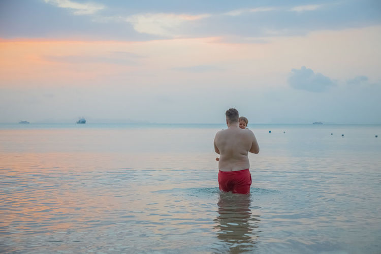 Rear view of man standing in sea against sky during sunset