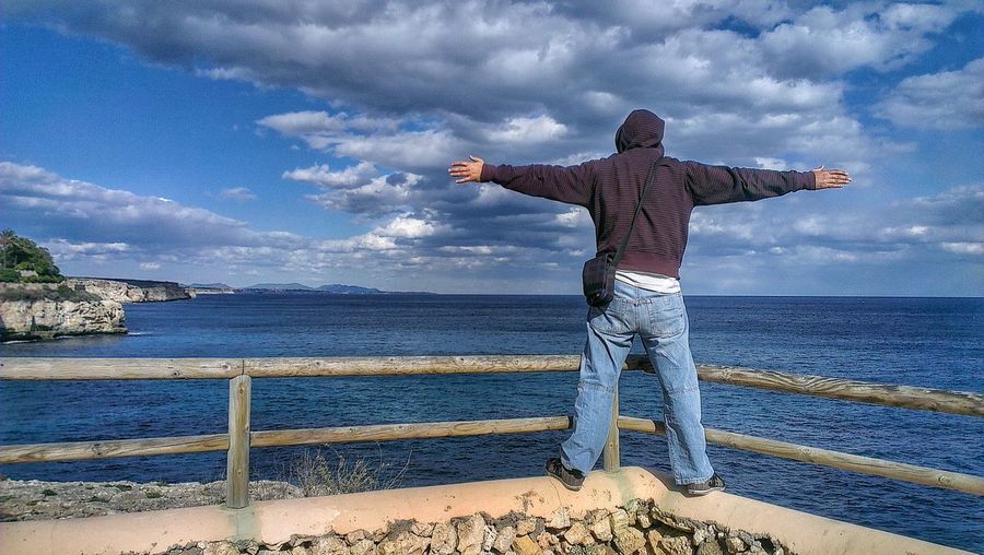 Rear view of man with arms outstretched standing against sea and cloudy sky