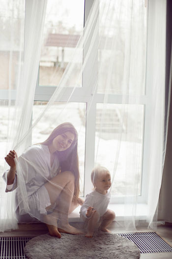 Mother in a white robe sits with a child a blonde daughter at a large window of the house person