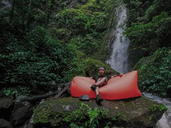 Man sitting on inflatable sofa against waterfall in forest