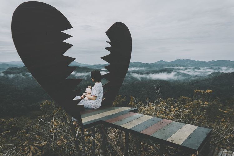 Teenage girl sitting on built structure with broken heart shape against mountain range