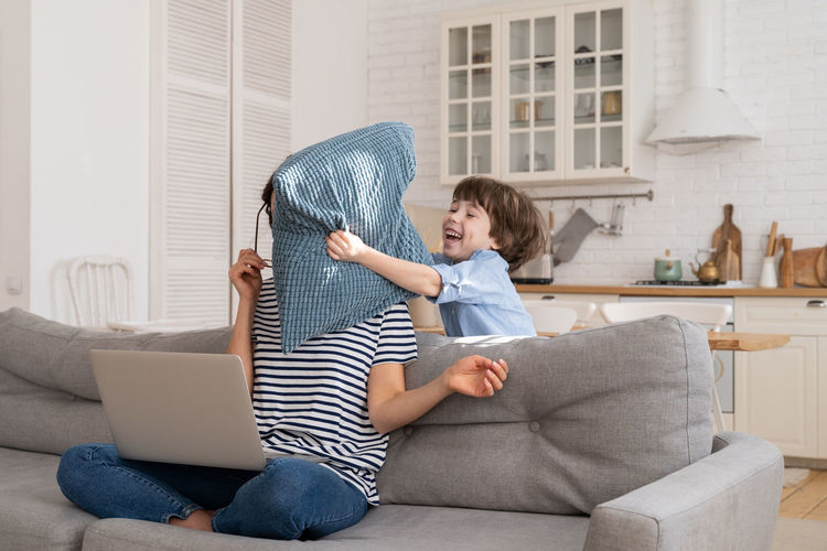 Exhausted mother sitting on couch at home work on laptop, child distracts, attacking mom with pillow